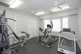 This fitness center offers a comprehensive workout environment housed in a single, thoughtfully arranged room. The space features light blue-gray walls, white trim, and a durable dark rubber flooring that provides cushioning and traction underfoot. Multiple windows dressed with white horizontal blinds allow natural light to filter in while maintaining privacy, contributing to an open and airy atmosphere.

The room is equipped with a variety of cardio machines, including a Matrix treadmill, an elliptical trainer, a recumbent bike, and an upright stationary bike — offering users a well-rounded selection of low- and high-impact cardiovascular options. A multi-function cable and weight machine anchors one side of the room, providing a range of strength training exercises in a compact footprint.

Two ceiling fans circulate air throughout the space, complemented by overhead fluorescent lighting that ensures the room remains well-lit at all hours. A wall-mounted flat-screen television provides entertainment during workouts, and a fitness center rules and information sign is posted prominently on the wall. The room also includes a door that opens to an adjacent area or exterior. Overall, the space is clean, functional, and designed to support a variety of fitness routines in an accessible, comfortable setting.