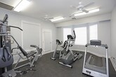 This fitness center offers a comprehensive workout environment housed in a single, thoughtfully arranged room. The space features light blue-gray walls, white trim, and a durable dark rubber flooring that provides cushioning and traction underfoot. Multiple windows dressed with white horizontal blinds allow natural light to filter in while maintaining privacy, contributing to an open and airy atmosphere.

The room is equipped with a variety of cardio machines, including a Matrix treadmill, an elliptical trainer, a recumbent bike, and an upright stationary bike — offering users a well-rounded selection of low- and high-impact cardiovascular options. A multi-function cable and weight machine anchors one side of the room, providing a range of strength training exercises in a compact footprint.

Two ceiling fans circulate air throughout the space, complemented by overhead fluorescent lighting that ensures the room remains well-lit at all hours. A wall-mounted flat-screen television provides entertainment during workouts, and a fitness center rules and information sign is posted prominently on the wall. The room also includes a door that opens to an adjacent area or exterior. Overall, the space is clean, functional, and designed to support a variety of fitness routines in an accessible, comfortable setting.