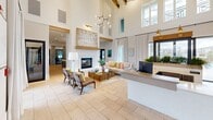 Located in Youngsville, Louisiana, this single-floor community clubhouse offers a generously sized, thoughtfully designed gathering space that blends modern aesthetics with warm, inviting details. The grand entrance and reception area immediately impress with soaring double-height ceilings, exposed wood beam trusses, and large clerestory windows that flood the space with natural light. Light travertine tile flooring flows throughout, unifying each area seamlessly.

The expansive living room anchors the space with a striking floor-to-ceiling white brick fireplace, a contemporary brass chandelier, and comfortable seating arrangements that invite relaxed conversation. Large sliding glass doors and oversized windows frame outdoor views and allow natural light to pour in from multiple directions.

The open-concept dining area features high-top tables with woven rattan chairs and direct access to an outdoor patio, creating a smooth indoor-outdoor connection. A well-appointed kitchen showcases white shaker cabinetry, subway tile backsplash, dark lower cabinets with brushed gold hardware, quartz countertops, a central island, and globe pendant lights in a warm brass finish — a functional yet stylish space for entertaining.

Throughout, curated artwork, lush indoor plants, and carefully selected furnishings in neutral tones with coastal accents contribute to a polished, welcoming ambiance ideal for community gatherings.