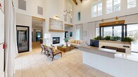 Located in Youngsville, Louisiana, this single-floor community clubhouse offers a generously sized, thoughtfully designed gathering space that blends modern aesthetics with warm, inviting details. The grand entrance and reception area immediately impress with soaring double-height ceilings, exposed wood beam trusses, and large clerestory windows that flood the space with natural light. Light travertine tile flooring flows throughout, unifying each area seamlessly.

The expansive living room anchors the space with a striking floor-to-ceiling white brick fireplace, a contemporary brass chandelier, and comfortable seating arrangements that invite relaxed conversation. Large sliding glass doors and oversized windows frame outdoor views and allow natural light to pour in from multiple directions.

The open-concept dining area features high-top tables with woven rattan chairs and direct access to an outdoor patio, creating a smooth indoor-outdoor connection. A well-appointed kitchen showcases white shaker cabinetry, subway tile backsplash, dark lower cabinets with brushed gold hardware, quartz countertops, a central island, and globe pendant lights in a warm brass finish — a functional yet stylish space for entertaining.

Throughout, curated artwork, lush indoor plants, and carefully selected furnishings in neutral tones with coastal accents contribute to a polished, welcoming ambiance ideal for community gatherings.