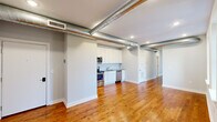 This thoughtfully designed unit offers five bedrooms and two bathrooms across a single floor, blending industrial character with modern finishes. Exposed silver ductwork runs throughout the space, creating a distinctive loft-inspired aesthetic complemented by crisp white walls and rich hardwood flooring that flows seamlessly from room to room.

The open-concept kitchen is outfitted with stainless steel appliances, including a refrigerator, range, microwave, and dishwasher, all set against white cabinetry and a classic subway tile backsplash. The kitchen opens directly to a generous common area, making it ideal for shared living.

The bedrooms vary in size, with the primary bedroom offering a comfortable footprint and natural light through double-hung windows framing views of the surrounding neighborhood. Additional bedrooms are equally well-lit, with some featuring exposed ductwork details consistent with the unit's industrial design language.

Both bathrooms are finished with white vanities, large mirrors, and standout decorative tile flooring in a bold blue-and-white medallion pattern, adding an elegant contrast to the otherwise neutral palette. A hallway connects the bedrooms and bathrooms, ensuring a functional and organized layout throughout this well-appointed multi-bedroom unit.