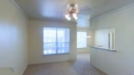 This single-floor residence offers a welcoming, open-concept layout that maximizes both space and natural light. The living area is bathed in natural light streaming through large windows fitted with horizontal blinds, creating a bright and airy atmosphere throughout the day. Soft, neutral-toned carpeting anchors the living space, lending warmth and comfort underfoot.

A ceiling fan with a multi-bulb light fixture serves as a functional centerpiece in the living area, providing both illumination and ventilation. The open layout transitions smoothly into a dining area, where an elegant pendant chandelier hangs overhead, adding a refined touch to the space.

The kitchen is partially open to the main living area and features a raised breakfast bar counter, ideal for casual dining or entertaining. Upper cabinetry in a light finish lines the kitchen walls, offering ample storage while maintaining the home's clean, cohesive aesthetic.

Crown molding details along the upper walls add an architectural accent that elevates the overall design. Multiple windows throughout the space ensure consistent natural light from various angles, contributing to the home's open and inviting ambiance. The neutral color palette used throughout creates a versatile backdrop suited to a wide range of interior styles.