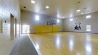 This indoor basketball facility offers a well-designed athletic space built for serious play and recreation alike. The centerpiece is a polished hardwood court, finished to a high sheen and marked with regulation court lines, providing excellent traction and performance underfoot. Towering ceilings give the space a generous vertical clearance, accommodating full gameplay without restriction. A regulation glass backboard and hoop are mounted along the far wall, which is lined with padded yellow safety panels to protect players near the boundary. Recessed flush-mount lighting is distributed evenly across the ceiling, ensuring consistent, shadow-free illumination throughout the court. A row of high, horizontally oriented windows runs along the upper portion of the walls, supplementing the artificial lighting with natural daylight. A transition mat at the court entrance helps maintain the floor surface. The neutral gray walls and clean architectural lines give the space a modern, functional aesthetic. Double glass doors provide convenient access from an adjacent area. Athletic wall art adds a dynamic visual accent. Overall, the space is purposefully designed to deliver a high-quality indoor sports experience in a clean, well-maintained environment.
