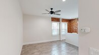 Located at 1057 S Western Ave in Los Angeles, this single-floor unit blends classic architectural character with fresh, modern finishes. The main living area makes an immediate impression with striking exposed red brick walls that wrap around multiple corners, creating a warm, industrial-chic aesthetic. Large windows fitted with white blinds allow natural light to fill the space, complementing the light-toned wood-look flooring that runs throughout. A sleek black ceiling fan with integrated lighting anchors the living area, enhancing both comfort and style. Recessed lighting adds a clean, contemporary touch to the ceilings. A low white partition wall defines the kitchen area, which is equipped with stainless steel appliances and benefits from abundant window light framed by the signature brick walls. The bedroom is a bright, neutral retreat with clean white walls, wood-look flooring, and access to a dedicated walk-in closet complete with built-in shelving. An additional storage space is also present. The bathroom features a full tub and shower combo with white tile surround, a vanity with cabinet storage, and wood-look flooring, maintaining the unit's consistent, clean aesthetic throughout.