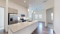 This well-designed single-floor residence offers a bright, open-concept layout that seamlessly connects the kitchen and living areas. The kitchen is a standout feature, showcasing a large central island with granite countertops, an undermount sink with a brushed gold faucet, and open shelving at the base. White and grey cabinetry lines the walls, complemented by a classic tile backsplash and a full suite of stainless steel appliances including a refrigerator, electric range, microwave, and dishwasher. Pendant lighting and recessed fixtures illuminate the space, which benefits from soaring ceilings that enhance the sense of openness. The living area continues the airy feel with tall ceilings, a ceiling fan, wood-style plank flooring, and multiple windows and a glass door that invite abundant natural light. Bedrooms feature plush carpet flooring, tall ceilings, and natural light from windowed walls. The bathroom is finished with large-format neutral tile surrounding a soaking tub and shower combination, accented by brushed nickel fixtures. A dedicated laundry room with overhead shelving and a washer/dryer unit adds everyday convenience. The consistent use of neutral tones, modern finishes, and thoughtful design creates a clean and functional living environment throughout.
