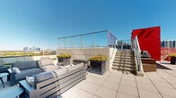 Located in Chicago's vibrant Old Town neighborhood, this rooftop amenity level at 1225 N Wells St offers an exceptional outdoor experience set against a striking backdrop of the city skyline. The space is defined by its modern architectural aesthetic, featuring sleek glass railings, clean concrete surfaces, and bold design elements including a vivid red structural facade that adds a dynamic visual contrast to the surrounding cityscape.

The rooftop is organized into several distinct functional zones. A resort-style pool serves as a central focal point, accessed via glass-paneled staircases that maintain unobstructed sightlines across the deck. Multiple open-air seating areas are thoughtfully arranged throughout the space, outfitted with contemporary outdoor furniture ideal for lounging or dining. A dedicated grilling station with a full-size gas grill and wooden prep surface provides a well-equipped outdoor cooking area.

Elevated walkways with glass and metal railings connect different sections of the rooftop, offering sweeping panoramic views of the Chicago skyline, including prominent downtown towers to the south. Planters with seasonal greenery add natural elements throughout the deck. The building's distinctive blue glass curtain wall rises adjacent to the rooftop, reinforcing the space's contemporary architectural character. This amenity level delivers a compelling urban outdoor retreat with broad, unobstructed city views in every direction.