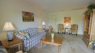 This single-floor space presents a comfortable and well-arranged living area that emphasizes a classic, homey aesthetic. The main sitting area is anchored by a blue and white checkered sofa paired with two striped wingback chairs, creating an inviting conversational arrangement around a wooden coffee table adorned with a floral centerpiece. Neutral-toned carpeting runs throughout, lending warmth and continuity to the space. The walls are painted in a soft, light hue that enhances the room's brightness. Decorative artwork adds personality to the room, including a vibrant autumn landscape painting and a bold floral print, both contributing color and visual interest. Soft ambient lighting is provided by table lamps positioned throughout the seating area, creating a warm atmosphere. The layout is practical and open, with a clearly visible entry door providing a natural focal point and easy access. Overall, the space conveys a relaxed, traditional character well-suited for comfortable everyday living.