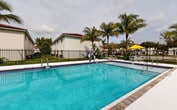 This Florida City property boasts a striking centerpiece: a large, inviting swimming pool with crystal-clear turquoise water. The pool area is meticulously designed with a wide, clean concrete deck perfect for lounging. Safety features include sturdy metal railings and clear depth markers. Surrounding the pool, a black metal fence provides security while maintaining an open feel.

The landscape is adorned with mature palm trees, creating a tropical oasis ambiance. Lush greenery, including various tropical plants, enhances the natural beauty of the space. For shade and relaxation, yellow umbrellas and comfortable lounge chairs are strategically placed around the pool.

The property's architecture features white stucco buildings with terracotta-tiled roofs, typical of Mediterranean-inspired Florida design. These two-story structures offer a clean, modern aesthetic that complements the natural surroundings. The overall design creates a harmonious blend of built and natural elements, providing residents with a serene and inviting outdoor living space reminiscent of a high-end resort.