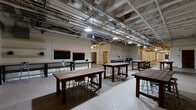 Located at 215 N First Ave E in Duluth, Minnesota, this single-floor space offers a generously sized, open-plan interior that blends industrial character with functional design. The layout is defined by exposed ceiling infrastructure — including ductwork, conduit, and piping — suspended above polished and terrazzo concrete floors, creating a raw yet purposeful aesthetic throughout.

The main open area is furnished with a series of sturdy, rustic wood communal tables paired with industrial-style stools, arranged to encourage collaboration and flexibility. Pendant lighting fixtures hang from the ceiling structure, providing warm, focused illumination across the workspace. Blackboards and wall-mounted panels are visible along one section, suggesting utility for presentations or instructional use.

A corridor extends through the space, flanked by painted white brick walls that speak to the building's historic origins. Arched brick detailing frames interior doorways, reinforcing the character of the original architecture. Utility sinks and work benches are present in a dedicated section, pointing to hands-on or maker-oriented uses.

Framed artwork and blueprint prints decorate select walls, adding a curated touch to the otherwise utilitarian environment. Multiple doorways — including classic wood-paneled doors — connect to adjacent rooms and ancillary areas. The overall ambiance is that of a creative, industrial-style collaborative or educational facility with a strong sense of heritage and openness.