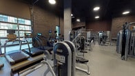 Located at 1245 W Fulton Market in Chicago's vibrant Fulton Market district, this single-floor fitness facility combines industrial aesthetics with a thoroughly equipped workout environment. The space is characterized by exposed brick walls, dramatic high ceilings, and dark matte finishes that create a sleek, urban atmosphere throughout.

Large glass garage-style doors line one wall, flooding the cardio zone with natural light and offering views of the surrounding streetscape. This area houses rowing machines, stationary bikes, and elliptical trainers. Overhead, recessed circular lighting supplements natural light across the facility.

The strength training area is expansive, featuring a wide selection of selectorized machines, cable systems, and free-weight stations. A dedicated free weights section includes a full rack of dumbbells, adjustable benches, and barbell setups with weight plate storage. Full-length mirrors are strategically positioned throughout to assist with form and expand the sense of space.

A functional training zone includes battle ropes, TRX suspension systems, kettlebells, and step platforms, supported by wall-mounted displays. Concrete support columns punctuate the floor plan, reinforcing the building's industrial character. The flooring transitions between rubber fitness flooring and hardwood in select areas, defining different workout zones while maintaining a cohesive, polished aesthetic.
