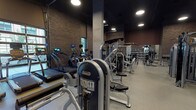 Located at 1245 W Fulton Market in Chicago's vibrant Fulton Market district, this single-floor fitness facility combines industrial aesthetics with a thoroughly equipped workout environment. The space is characterized by exposed brick walls, dramatic high ceilings, and dark matte finishes that create a sleek, urban atmosphere throughout.

Large glass garage-style doors line one wall, flooding the cardio zone with natural light and offering views of the surrounding streetscape. This area houses rowing machines, stationary bikes, and elliptical trainers. Overhead, recessed circular lighting supplements natural light across the facility.

The strength training area is expansive, featuring a wide selection of selectorized machines, cable systems, and free-weight stations. A dedicated free weights section includes a full rack of dumbbells, adjustable benches, and barbell setups with weight plate storage. Full-length mirrors are strategically positioned throughout to assist with form and expand the sense of space.

A functional training zone includes battle ropes, TRX suspension systems, kettlebells, and step platforms, supported by wall-mounted displays. Concrete support columns punctuate the floor plan, reinforcing the building's industrial character. The flooring transitions between rubber fitness flooring and hardwood in select areas, defining different workout zones while maintaining a cohesive, polished aesthetic.
