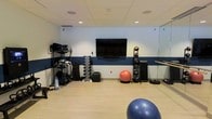 This single-level fitness space is designed with functionality and versatility in mind. The room features a clean, open floor plan with light wood-style flooring that provides a comfortable, durable surface suitable for a range of exercises. The drop ceiling with recessed lighting ensures the space is well-lit, creating a bright and energizing workout environment.

A bold navy blue accent stripe runs along the perimeter walls, adding a dynamic visual element against the neutral upper walls. A large wall-mounted flat-screen TV is centrally positioned, allowing users to follow workout videos or entertainment during exercise sessions.

The space is outfitted with an impressive array of equipment, including a full dumbbell rack with multiple weight increments, a medicine ball tower, stability balls in varying sizes, weight plates, and additional accessories stored on a multi-tier rack. A wall-mounted ballet barre and a mirrored wall panel on one side add a studio-like quality, making the space adaptable for stretching, balance work, or dance-based fitness activities. Overall, the space is practical, organized, and comprehensively equipped.