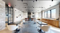 This single-floor space showcases a refined industrial-contemporary aesthetic, combining raw materials with sleek modern furnishings to create a highly functional and visually compelling environment. Polished concrete floors span the entire open-plan layout, reflecting natural light that pours in through large, black-framed windows offering views of the surrounding urban landscape. Exposed brick columns punctuate the interior, adding textural contrast against crisp white walls adorned with small wall-mounted display shelves.

The ceiling features a dark-painted finish fitted with track lighting, pendant lights, and cylindrical hanging fixtures, providing layered and adjustable illumination throughout. Workstations include dark-topped communal tables paired with modern white molded chairs on casters, as well as a central bar-height table surrounded by warm-toned wooden stools. A lounge area near the windows offers a cognac leather sofa and accent seating for informal gatherings.

Steel-framed glass partition walls divide a separate enclosed room, visible from the main area, maintaining an open and transparent feel while providing a degree of privacy. Warm wood cabinetry lines one windowed wall, adding a natural, grounding element to the otherwise industrial palette. The overall ambiance is clean, bright, and professionally inviting.