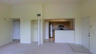 This single-story space is characterized by its notably high ceilings, which lend an airy, open quality to the interior. The main living area features neutral-toned carpet and tall walls painted in a soft cream, creating a clean and versatile backdrop. A built-in closet with double rods and shelving is conveniently situated adjacent to the living space, offering practical storage.

The kitchen is positioned behind a raised pass-through counter that separates it from the living area while maintaining an open connection between the two spaces. The kitchen is equipped with stainless steel appliances, including a refrigerator, microwave, and range, complemented by warm-toned wood cabinetry that extends to the upper walls. Light tile flooring in the kitchen contrasts with the carpeted living area, clearly defining each zone.

Overhead, a ceiling vent is visible, indicating a central HVAC system. The overall design is functional and straightforward, with an emphasis on maximizing vertical space and maintaining a cohesive, neutral aesthetic throughout.