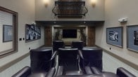 This elegantly designed media lounge combines dramatic architectural elements with practical amenities, creating a refined gathering space ideal for entertainment and work alike. The room commands attention with its soaring double-height ceilings, which are accentuated by a striking ornamental wrought-iron balcony with rich wood detailing — a standout architectural feature that adds a classic, upscale character to the space.

The main seating area is furnished with a generous arrangement of dark leather club chairs, oriented toward a large flat-screen television recessed into a niche flanked by rustic wood doors and tall potted plants, creating a focused, theater-like viewing experience. Recessed lighting and decorative wall sconces provide warm, ambient illumination throughout.

A collection of black-and-white framed artwork adorns the walls, lending a curated, gallery-like atmosphere. Adjacent to the main lounge area, a built-in workstation with a clean white desk surface and upholstered chairs offers a quiet area for productivity. Wood-framed windows with slatted blinds allow natural light to filter in while maintaining privacy.

The soft, neutral carpet and warm taupe walls tie the entire space together, creating an inviting and cohesive environment that balances comfort, style, and function.