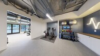 This fitness center in Berkeley, CA (94704) is a thoughtfully designed, single-floor facility that blends industrial aesthetics with functional workout spaces. The open layout features exposed ceiling infrastructure with visible ductwork and piping painted in dark tones, complemented by bright linear and pendant lighting that ensures excellent visibility throughout.

The main cardio and strength area is naturally lit by large street-facing windows and glass doors, allowing abundant natural light and views of the surrounding neighborhood. This section houses multiple treadmills, a power rack with weight bench, free weights, and additional strength equipment arranged to maximize floor space.

An adjoining cycling area features two monitor-equipped stationary bikes positioned on protective floor mats against a textured blue accent wall. Nearby, a well-organized equipment rack displays a full range of kettlebells, medicine balls, resistance tools, and stability balls for varied training options.

A separate open studio room provides a clean, minimalist space with rubberized flooring, large wall mirrors, and a mirror-mounted interactive fitness display — ideal for stretching, yoga, or guided workouts. Restroom facilities are conveniently accessible within the space, marked with clear signage. Throughout, the durable speckled flooring and white walls maintain a clean, professional atmosphere suited for diverse fitness activities.