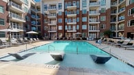Located at 1108 S Main St in Greenville, South Carolina, this outdoor amenity space is designed to offer a resort-style experience within a modern residential community. The centerpiece of the space is a generously sized swimming pool with clear, blue water, complemented by an adjacent spa. The pool deck is finished in large-format stone tiles, providing an elegant and durable surface throughout the courtyard. Surrounding the pool, an array of lounge chairs and umbrella-shaded seating invites residents to relax in the open air.

The covered outdoor kitchen and bar area is a standout feature, equipped with expansive granite countertops, a built-in sink with a professional-style faucet, and a wall-mounted flat-screen television. Brick accent walls and recessed lighting add character and warmth to this covered entertainment space, making it well-suited for gatherings.

Beyond the pool area, a dedicated lounge zone offers comfortable wicker and upholstered seating arrangements set against the backdrop of the building's classic brick façade with large windows and private balconies. Mature plantings and ornamental trees are thoughtfully integrated throughout the courtyard, enhancing the overall ambiance. The multi-story brick exterior frames the entire space, creating an enclosed, community-oriented atmosphere in the heart of Greenville.