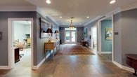 This single-floor space presents a well-appointed interior with a traditional aesthetic throughout. The main foyer serves as the heart of the layout, featuring large slate tile flooring, crown molding, and a decorative lantern-style pendant light that anchors the entryway. French doors with multi-pane glass panels flood the space with natural light and provide a view of the surrounding greenery outside.

The foyer transitions seamlessly into a comfortable lounge area furnished with a tufted Chesterfield sofa, classic wingback chairs, and a wooden bookcase, all arranged atop an ornate area rug. The muted blue-gray walls provide a calm, cohesive backdrop throughout, complemented by white trim and recessed lighting overhead.

Adjacent areas include a teal-accented mail room with a corkboard, a utility cart, and organized storage, as well as a private office visible through a doorway. Informational displays are mounted along the walls, and a full-length mirror expands the sense of space in the lounge. Traditional wooden accent furniture, table lamps, and artwork contribute to the refined, welcoming character of this well-organized communal environment.