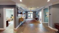 This single-floor space presents a well-appointed interior with a traditional aesthetic throughout. The main foyer serves as the heart of the layout, featuring large slate tile flooring, crown molding, and a decorative lantern-style pendant light that anchors the entryway. French doors with multi-pane glass panels flood the space with natural light and provide a view of the surrounding greenery outside.

The foyer transitions seamlessly into a comfortable lounge area furnished with a tufted Chesterfield sofa, classic wingback chairs, and a wooden bookcase, all arranged atop an ornate area rug. The muted blue-gray walls provide a calm, cohesive backdrop throughout, complemented by white trim and recessed lighting overhead.

Adjacent areas include a teal-accented mail room with a corkboard, a utility cart, and organized storage, as well as a private office visible through a doorway. Informational displays are mounted along the walls, and a full-length mirror expands the sense of space in the lounge. Traditional wooden accent furniture, table lamps, and artwork contribute to the refined, welcoming character of this well-organized communal environment.