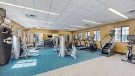 Located at 2701 Fletcher Ave. in Lincoln, Nebraska, this fitness center offers a comprehensive workout environment within a well-organized, single-floor space. The room features warm golden-yellow walls, a drop-panel ceiling with integrated linear lighting, and a combination of teal patterned carpet and light geometric-tiled flooring that visually defines different workout zones.

The cardio zone is equipped with multiple treadmills, stair climbers, and a stationary cycling bike, each outfitted with individual display screens. The strength training area includes a multi-station cable machine, a leg press, a power rack with weight plates, and a dedicated dumbbell rack with wall-mounted mirrors that enhance the sense of space and allow for form monitoring. A functional training corner rounds out the facility, featuring a multi-purpose rig, battle ropes, medicine balls in various sizes, a heavy bag, and resistance accessories.

Multiple windows line the walls, allowing natural light to fill the space and offering views to the surrounding outdoor areas. The layout is thoughtfully arranged to accommodate a variety of fitness activities simultaneously without crowding. A water fountain is also present near the entrance area, adding a convenient amenity for users. Overall, this fitness center provides a fully equipped, bright, and functional environment suited for a wide range of exercise routines.