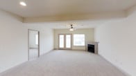 This single-floor space presents an open and airy living area designed for comfort and functionality. The main living space is generously sized, featuring plush wall-to-wall carpeting in a neutral tone that contributes to the room's warm, inviting atmosphere. A ceiling fan with a light fixture is centrally positioned, providing both air circulation and ambient lighting.

A tile-surround fireplace anchors one wall of the living area, serving as a natural focal point and adding a cozy architectural element to the space. Adjacent to the fireplace, a large window and a set of French doors flood the room with natural light while offering direct access to the outdoors. The French doors and window together create a bright, open feel that enhances the sense of spaciousness.

The neutral, cream-toned walls and ceiling complement the light carpeting, creating a cohesive and understated palette throughout. A wide doorway connects the main living area to an adjoining room, also carpeted, allowing for a seamless flow between spaces. Overall, the layout is practical and versatile, well-suited for a variety of living arrangements.