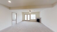 This single-floor space presents an open and airy living area designed for comfort and functionality. The main living space is generously sized, featuring plush wall-to-wall carpeting in a neutral tone that contributes to the room's warm, inviting atmosphere. A ceiling fan with a light fixture is centrally positioned, providing both air circulation and ambient lighting.

A tile-surround fireplace anchors one wall of the living area, serving as a natural focal point and adding a cozy architectural element to the space. Adjacent to the fireplace, a large window and a set of French doors flood the room with natural light while offering direct access to the outdoors. The French doors and window together create a bright, open feel that enhances the sense of spaciousness.

The neutral, cream-toned walls and ceiling complement the light carpeting, creating a cohesive and understated palette throughout. A wide doorway connects the main living area to an adjoining room, also carpeted, allowing for a seamless flow between spaces. Overall, the layout is practical and versatile, well-suited for a variety of living arrangements.