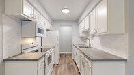 This single-floor residence presents a clean, updated aesthetic throughout its well-organized layout. The galley-style kitchen is a standout feature, offering ample white shaker cabinetry with brushed nickel hardware on both sides, gray laminate countertops, a classic white subway tile backsplash, and a stainless steel sink. White appliances, including an electric range, built-in microwave, and refrigerator, are neatly integrated. Wood-look plank flooring runs through the kitchen and living areas, adding warmth and continuity. The living room is open and inviting, featuring a ceiling fan, sliding glass door that opens to a green outdoor view, and generous wall space. Bedrooms are finished with plush carpet and neutral gray walls, with multiple windows allowing natural light. The bathroom features wood-look flooring, a white vanity with a gray countertop and framed mirror, and a full tub-shower combination enclosed by a sleek sliding glass panel and surrounded by crisp white subway tile. An electrical panel is conveniently located along the interior hallway. The overall palette of soft grays and whites creates a cohesive, modern feel throughout.