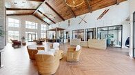 Located at 2175 Easton Loop in Kissimmee, Florida, this expansive single-floor clubhouse delivers an impressive resort-style experience through thoughtful design and high-end finishes. The grand entrance lobby sets the tone immediately, showcasing soaring vaulted ceilings with rich wood detailing, dramatic floor-to-ceiling windows, and herringbone-patterned wood-look tile flooring that flows throughout the main living areas. A prominent reception desk anchors the lobby, flanked by comfortable wicker seating and abundant natural light from large black-framed windows overlooking the outdoors. The main great room is a showstopper, featuring a cathedral ceiling with exposed wood beams, a statement chandelier, a stone-clad fireplace, and an open layout that seamlessly connects lounge, dining, and bar zones. A stylish bar area features bold blue cabinetry, tiled accents, pendant lighting, and a custom wood-and-metal shelving partition. Throughout the space, built-in bookshelves serve as elegant room dividers, adding warmth and character to lounge corridors lined with curated artwork. A dedicated game room stands out with vibrant teal walls, geometric-patterned tile flooring, a shuffleboard table, arcade games, a wall-mounted TV, and large windows with pool and community views. The overall ambiance is contemporary coastal — warm, inviting, and thoughtfully designed for community living.