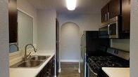 This single-floor space showcases a well-appointed kitchen that serves as a central highlight of the layout. The kitchen is equipped with dark espresso-toned cabinetry that provides ample storage, complemented by a clean white countertop and a double-basin stainless steel sink. Stainless steel appliances, including a refrigerator, a gas range with black finish, and an over-the-range microwave, add a modern and functional character to the space. A flush-mounted ceiling light illuminates the kitchen efficiently, enhancing the clean, neutral gray walls. One of the most distinctive architectural features is an elegant arched passageway that adds a refined, classic touch to the interior design, creating a graceful transition between spaces. The flooring consists of rich, dark hardwood-style planks that run throughout, anchoring the space with warmth and continuity. The overall ambiance is practical yet stylish, with a cohesive design palette of dark cabinetry, neutral walls, and dark flooring that creates a balanced and inviting environment. The thoughtful layout maximizes functionality while maintaining a polished, contemporary aesthetic throughout.