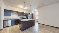 This single-floor unit showcases a thoughtfully designed open-concept layout that seamlessly connects the kitchen and living areas. The kitchen serves as the centerpiece, featuring rich dark cabinetry paired with granite countertops that extend across both the perimeter counters and a central island. Stainless steel appliances, including a refrigerator, electric range, and microwave, provide a sleek, modern finish. Pendant lights hang above the island, complementing a track lighting fixture overhead, while wood-style luxury flooring runs throughout the main living areas, adding warmth and continuity.

The living area is bright and open, benefiting from natural light through a window with blinds, and is equipped with a ceiling fan. A decorative wine rack built into the upper cabinetry adds a distinctive design touch to the kitchen.

The bedroom features plush carpeting and large windows that allow ample natural light to filter in. The bathroom is finished consistently with the rest of the unit, offering a granite-topped vanity with dark cabinetry, a large mirror, and a full tub-shower combination with grab bar. The overall color palette of neutral walls, dark trim, and wood-tone floors creates a cohesive, contemporary ambiance throughout.