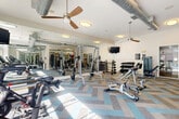 This fitness center is a thoughtfully designed, single-floor exercise facility offering a comprehensive workout environment. The space is generously sized, providing ample room for a wide variety of equipment and free movement throughout. A striking herringbone-patterned carpet in neutral tones accented with blue adds a modern, stylish touch underfoot.

Natural light pours in through large floor-to-ceiling windows along the perimeter, offering views of a nearby outdoor pool and lush greenery, creating an energizing and open atmosphere. The high ceilings, adorned with exposed industrial-style silver ductwork, add architectural character, while ceiling fans help maintain airflow throughout the space.

The cardio zone is well-stocked with a row of treadmills, elliptical machines, and stationary cycling bikes, each equipped with individual monitor screens. A wall-mounted television provides additional entertainment for users during workouts. The strength training area features a robust selection of cable machines, multi-functional weight stations, adjustable benches, and free weights. Floor-to-ceiling mirrors line one wall, visually expanding the space and allowing users to monitor their form. Structural columns are integrated seamlessly into the layout without disrupting the flow of the space. Overall, the fitness center combines functionality with a clean, contemporary aesthetic.