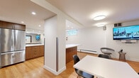 This well-appointed single-floor space offers a thoughtfully designed open-plan layout that seamlessly integrates multiple functional zones. The warm hardwood flooring runs continuously throughout, unifying the space and contributing to its inviting, contemporary atmosphere.

The kitchen area is a standout feature, equipped with sleek walnut-toned cabinetry, a generous white quartz countertop island, a stainless steel double sink with a modern pull-down faucet, and a full-size stainless steel refrigerator. Recessed lighting illuminates the kitchen, enhancing its clean, functional aesthetic.

The main lounge and gathering area is furnished with multiple grey sofas arranged around white dining tables and modern black chairs, making it well-suited for both social gatherings and casual work sessions. Wall-mounted flat-screen TVs add to the utility of the space. A large mirror accent wall visually expands the room, amplifying the sense of openness.

Natural light filters in through above-grade horizontal windows positioned along the upper walls, complemented by flush circular ceiling light fixtures and recessed lighting. The space is accessed through a black-framed glass-panel door, and includes practical amenities such as baseboard heating, a fire extinguisher, and a wall-mounted waste disposal unit.