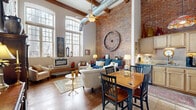 Located at 79 Warren St in Glens Falls, NY, this distinctive single-floor residence blends industrial character with warm, inviting design. The open-concept living and dining area is the heart of the home, boasting dramatically high ceilings with exposed wooden beams and a prominent ductwork detail that emphasizes its loft-like aesthetic. A full-height exposed brick wall serves as a stunning focal point, complemented by large arched windows that flood the space with natural light. Rich hardwood floors run throughout the living area, adding warmth and continuity. The kitchen features light wood cabinetry, stone countertops, stainless steel appliances, and tile flooring, finished with a colorful stained-glass pendant light. The spacious bedroom offers soaring ceilings, plush carpet, a generous closet, and a window with natural light. A dedicated office provides a quiet retreat with its own high ceilings and neutral tones. The bathroom is neatly appointed with a pedestal sink, tiled floors, and a shower. Each room reflects a thoughtful combination of architectural integrity and functional design, making this Glens Falls residence a truly unique and comfortable place to call home.