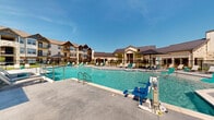Located at 10015 N Owasso Expy in Owasso, Oklahoma, this outdoor amenity area serves as a central gathering hub for the surrounding residential community. The centerpiece is a generously sized, resort-style swimming pool with crystal-clear turquoise water, complemented by a shallow wading ledge furnished with in-water lounge chairs — ideal for relaxing on warm Oklahoma days. An ADA-compliant pool lift ensures accessibility for all residents.

The expansive pool deck is finished in smooth concrete and lined with an array of sun loungers and umbrella-shaded seating, offering ample space for sunbathing and socializing. A covered pavilion, constructed with stone and brick accents, provides shaded outdoor seating and houses built-in grilling stations, making it well-suited for outdoor gatherings and barbecues.

The surrounding landscape features neatly manicured green lawns and ornamental plantings, enclosed by a black iron security fence. The architectural backdrop showcases multi-story craftsman-style residential buildings with board-and-batten siding, stone detailing, pitched gable rooflines, and private balconies. A clubhouse structure with a standing-seam metal roof and stone facade anchors one end of the pool area, adding a polished, upscale character to the overall outdoor environment.