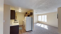 This single-floor space offers an open-concept design that seamlessly connects the kitchen and living area. The kitchen is equipped with dark wood cabinetry, a white refrigerator, a stainless steel oven, and a sink with a window view, all set on durable hardwood-style flooring. Upper and lower cabinetry provide ample storage, while light-toned countertops offer generous workspace. A textured popcorn ceiling runs throughout, adding a classic touch to the interior. The adjacent living area transitions to plush carpeting and is bathed in natural light, courtesy of a wide set of windows along the far wall that brighten the entire space. The warm, neutral wall tones throughout create a cohesive and welcoming ambiance. The open flow between the kitchen and living area makes the space feel expansive and versatile, suitable for both everyday living and casual entertaining. Overall, the layout is practical and well-connected, with functional finishes and a comfortable atmosphere.