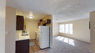 This single-floor space offers an open-concept design that seamlessly connects the kitchen and living area. The kitchen is equipped with dark wood cabinetry, a white refrigerator, a stainless steel oven, and a sink with a window view, all set on durable hardwood-style flooring. Upper and lower cabinetry provide ample storage, while light-toned countertops offer generous workspace. A textured popcorn ceiling runs throughout, adding a classic touch to the interior. The adjacent living area transitions to plush carpeting and is bathed in natural light, courtesy of a wide set of windows along the far wall that brighten the entire space. The warm, neutral wall tones throughout create a cohesive and welcoming ambiance. The open flow between the kitchen and living area makes the space feel expansive and versatile, suitable for both everyday living and casual entertaining. Overall, the layout is practical and well-connected, with functional finishes and a comfortable atmosphere.