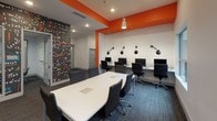 Located at 3165 E Michigan Ave in Lansing, Michigan, this single-floor amenity space is designed with a vibrant, modern aesthetic that blends functionality with an energetic atmosphere. Bold orange architectural beams and accent walls punctuate the bright white ceilings and walls, creating a dynamic visual identity throughout.

The main open area serves as a collaborative lounge and conference space, featuring a large white conference table surrounded by sleek black chairs, pendant lighting with industrial-style fixtures, and a vivid lime-green sofa paired with bold orange accent chairs. Multiple windows frame views of an outdoor terrace and the surrounding building exterior, filling the space with natural light.

A dedicated business center lines one wall with a row of desktop computer workstations, each equipped with a monitor and individual wall-mounted swing-arm task lamps. A striking chalkboard-style mural wall covered in mathematical equations and formulas adds an academic, creative energy to this area.

A smaller private meeting room offers a round wood-finish table, teal chairs, and a wall-mounted flat-screen television framed in red, accessible via glass-panel sliding doors. The space also includes a casual lounge nook with orange accent chairs and convenient waste and recycling stations near a glass exterior door. Blue-grey patterned carpet ties the entire space together cohesively.