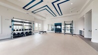 Located at 100 Windflower Ln in Daytona Beach, Florida, this fitness center offers a generously sized, single-floor exercise space designed to support a full range of workout routines. The room is bright and airy, bathed in natural light streaming through multiple large windows that frame views of the surrounding community. The crisp white walls and light-toned speckled flooring create a clean, energizing atmosphere throughout.

A standout architectural feature is the tray ceiling accented with bold navy blue geometric trim, adding a modern, design-forward aesthetic to the space. Ceiling fans are positioned throughout to maintain airflow and comfort during workouts. Recessed lighting provides consistent illumination across the entire floor.

The cardio zone is well-appointed with treadmills, elliptical machines, and stationary bikes arranged along the windowed walls. The strength training area includes multi-use power racks, a full dumbbell rack, medicine balls, and TRX suspension systems. A dedicated stretching and functional training area features padded flooring and adjustable benches, complemented by a wall of floor-to-ceiling mirrors that visually expand the space. A large flat-screen display is mounted within a navy blue accent wall, serving as a central focal point. Double glass entry doors provide a polished, welcoming entrance to this comprehensively equipped facility.
