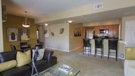 This single-floor residence offers an open-concept layout that seamlessly connects the living area to the kitchen, creating a spacious and cohesive environment. The living room is comfortably furnished with dark leather seating and accented by decorative wall art, including metallic leaf sculptures that add a contemporary touch to the warm, neutral-toned walls. A glass coffee table and sculptural décor contribute to the refined aesthetic of the space.

The kitchen is designed for both functionality and style, featuring stainless steel appliances, warm wood cabinetry, and a dedicated wine rack above the upper cabinets. A breakfast bar with tall black bar stools provides a casual dining option and serves as a natural dividing element between the kitchen and living areas. The dark countertops complement the cabinetry, giving the kitchen a polished, modern appearance.

Soft carpeting runs throughout the main living area, enhancing the warmth and comfort of the space. Recessed lighting in the ceiling, combined with flush-mount fixtures, ensures consistent and even illumination throughout. A window visible toward the far end of the space allows for the introduction of natural light. Overall, the space presents a well-balanced blend of contemporary design and everyday comfort.