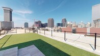 This amenity space combines an open-air rooftop dog run with a well-appointed fitness center, delivering a compelling mix of outdoor recreation and indoor wellness facilities. The rooftop dog run features lush artificial turf framed by sleek metal mesh fencing, providing a secure and comfortable environment for pets. The elevated position affords sweeping, panoramic views of an impressive urban skyline, with high-rise buildings stretching across the horizon beneath open blue skies — a remarkable backdrop for daily outdoor activity.

The fitness center is thoughtfully designed with clean, modern finishes including neutral walls, recessed lighting, and floor-to-ceiling mirrors that amplify the sense of space. Large glass doors and windows invite abundant natural light and offer views of the outdoor surroundings, maintaining an airy and energizing atmosphere. The gym is comprehensively equipped with treadmills, elliptical machines, and a full rack of dumbbells for strength training, alongside a multi-station cable machine, squat rack, barbells, weight plates, medicine balls, and an adjustable bench. Wooden floating shelves provide convenient storage for towels and accessories. A wall-mounted television adds entertainment during workouts, while a ceiling fan supports comfortable airflow throughout the space.
