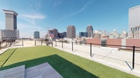 This amenity space combines an open-air rooftop dog run with a well-appointed fitness center, delivering a compelling mix of outdoor recreation and indoor wellness facilities. The rooftop dog run features lush artificial turf framed by sleek metal mesh fencing, providing a secure and comfortable environment for pets. The elevated position affords sweeping, panoramic views of an impressive urban skyline, with high-rise buildings stretching across the horizon beneath open blue skies — a remarkable backdrop for daily outdoor activity.

The fitness center is thoughtfully designed with clean, modern finishes including neutral walls, recessed lighting, and floor-to-ceiling mirrors that amplify the sense of space. Large glass doors and windows invite abundant natural light and offer views of the outdoor surroundings, maintaining an airy and energizing atmosphere. The gym is comprehensively equipped with treadmills, elliptical machines, and a full rack of dumbbells for strength training, alongside a multi-station cable machine, squat rack, barbells, weight plates, medicine balls, and an adjustable bench. Wooden floating shelves provide convenient storage for towels and accessories. A wall-mounted television adds entertainment during workouts, while a ceiling fan supports comfortable airflow throughout the space.