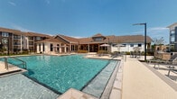 Located at 200 Mirage Ave in Crestview, Florida, this single-story outdoor amenity space delivers a resort-inspired experience designed for relaxation and recreation. At the heart of the space is a generously sized swimming pool with crystal-clear blue water, complemented by an adjacent wading pool or spa area, both finished with clean, contemporary hardscaping. The expansive pool deck is outfitted with lounge chairs, shaded umbrella tables, and seating areas that invite outdoor gatherings under the Florida sun. Pergola structures anchor multiple corners of the courtyard, providing shaded retreats with additional seating for socializing or unwinding. Lush, manicured green lawn areas border the pool zone, furnished with lounge chairs and accented by decorative plantings, mulched garden beds, and ornamental grasses that add texture and color throughout. A black metal perimeter fence provides a sense of enclosure while maintaining open sightlines to the surrounding multi-story residential buildings with private balconies. The overall layout, as revealed in the aerial view, forms a cohesive courtyard configuration with the clubhouse building wrapping the pool area and offering direct access to the outdoor space through large glass-panel doors and covered transitional areas.