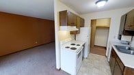 This single-floor residential space offers a practical and straightforward layout designed for comfortable everyday living. The kitchen serves as a central feature, equipped with a white electric coil range and a full-size refrigerator. Rich, dark wood upper cabinets provide ample storage, complemented by an under-cabinet range hood that keeps the cooking area ventilated. The kitchen flooring features light-toned vinyl tile, creating a clean and easy-to-maintain surface. Adjacent to the kitchen, the living area transitions to warm, wall-to-wall carpeting, offering a cozy and inviting atmosphere. The open connection between the kitchen and living space contributes to an efficient, flowing floor plan well-suited for daily use. A closet space is visible just off the kitchen, providing additional storage with a built-in shelf, ideal for pantry or organizational needs. The walls feature a combination of textures and warm tones, adding character to the interior. Overhead lighting in the kitchen and a textured ceiling throughout complete the functional aesthetic of this straightforward, well-appointed single-floor residence.