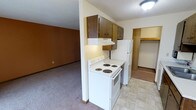 This single-floor residential space offers a practical and straightforward layout designed for comfortable everyday living. The kitchen serves as a central feature, equipped with a white electric coil range and a full-size refrigerator. Rich, dark wood upper cabinets provide ample storage, complemented by an under-cabinet range hood that keeps the cooking area ventilated. The kitchen flooring features light-toned vinyl tile, creating a clean and easy-to-maintain surface. Adjacent to the kitchen, the living area transitions to warm, wall-to-wall carpeting, offering a cozy and inviting atmosphere. The open connection between the kitchen and living space contributes to an efficient, flowing floor plan well-suited for daily use. A closet space is visible just off the kitchen, providing additional storage with a built-in shelf, ideal for pantry or organizational needs. The walls feature a combination of textures and warm tones, adding character to the interior. Overhead lighting in the kitchen and a textured ceiling throughout complete the functional aesthetic of this straightforward, well-appointed single-floor residence.