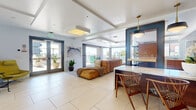 Located in San Diego's 92101 zip code, this single-floor clubhouse offers a generously proportioned communal space designed in a contemporary, mid-century modern aesthetic. Large-format tile flooring runs throughout the interior, reflecting abundant natural light that pours in through floor-to-ceiling windows and glass doors.

The open-plan living and dining area serves as the heart of the clubhouse. The living zone features built-in wall niches with mounted flat-screen televisions, structured ceiling details, and comfortable lounge seating. Multiple dining configurations are present, including a marble-topped island bar with sculptural mid-century wood chairs beneath brass pendant lights, a bold navy accent wall, and a separate dining table with tufted upholstered seating set atop a geometric area rug.

The kitchen is finished with warm wood cabinetry and stone countertops, while curated artwork and lush indoor plants add character throughout.

Sliding glass doors open onto a spacious outdoor terrace with glass-panel railings, offering sweeping views of the San Diego skyline and palm tree-lined cityscape. The terrace includes lounge seating, a rustic log-faced outdoor bar with counter seating, and café string lighting, creating an inviting alfresco setting.