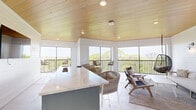 Located at 7950 Park Blvd N in Pinellas Park, Florida, this thoughtfully designed lounge space offers a seamless blend of modern coastal aesthetics and functional comfort. The single-floor layout is unified by a striking warm-toned wood-plank ceiling that adds natural texture and visual warmth throughout the space.

The living area is anchored by a comfortable neutral-toned sofa, woven accent chairs, and a central coffee table set atop a layered area rug. A pair of distinctive hanging egg chairs adds a playful, resort-inspired touch, suspended near large floor-to-ceiling windows that frame lush, elevated treetop views and open onto a railed balcony, flooding the interior with abundant natural light.

A well-appointed bar area serves as a central gathering point, featuring a long white quartz-topped island with bar seating, a wall-mounted television, and a built-in sink — making it ideal for entertaining. The surrounding shiplap-clad white walls complement the polished concrete flooring, creating a clean, contemporary backdrop throughout.

Stylish wall sconces provide warm accent lighting along the lounge wall, while recessed ceiling lights ensure the space is well-lit at all times. The open layout, expansive windows, and elevated outdoor vistas combine to create a bright, airy ambiance that is both inviting and versatile.