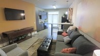 This single-floor space showcases a thoughtfully designed open-concept layout that seamlessly connects the living and kitchen areas into one cohesive, functional environment. The living room features a generously sized sectional sofa adorned with decorative throw pillows in warm earth tones, complemented by a tufted leather ottoman that serves as a stylish centerpiece. A wall-mounted flat-screen television and a rustic console table add both practicality and character to the space. The warm amber accent wall provides a bold contrast to the crisp white walls, creating a welcoming ambiance throughout. The kitchen is outfitted with sleek gray cabinetry, a dark countertop workspace, and black appliances, lending the area a modern and polished aesthetic. A breakfast bar with pendant lighting and wire bar stools creates a natural divide between the kitchen and living area, offering a casual dining or gathering spot. Light wood-look flooring runs throughout, tying the two spaces together with a clean, contemporary finish. Natural light filters in through a large window near the kitchen, brightening the interior and enhancing the open, airy feel of the overall layout.