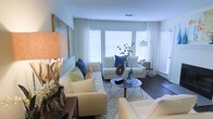 This single-floor space showcases a thoughtfully designed living area that emphasizes comfort and contemporary style. The living room is anchored by a light-toned sectional sofa and matching armchairs, accented with navy blue and soft green throw pillows that lend a coastal aesthetic to the room. A round, mid-century modern coffee table sits atop a patterned area rug, providing a focal point for the seating arrangement. The dark hardwood flooring offers a rich contrast to the predominantly white and cream color palette of the space. Large windows dressed with white horizontal blinds and flowing sheer curtains allow generous amounts of natural light to fill the room, creating an airy and open atmosphere. Decorative elements, including a sculptural table lamp with a natural wood base, abstract wall art in muted tones, and tasteful tabletop accessories in blue and neutral hues, contribute to the cohesive design scheme. An arc floor lamp adds additional ambient lighting. The overall ambiance is serene, modern, and welcoming, with careful attention to detail evident throughout the space.