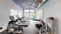 This fitness center is designed to accommodate a full range of workout needs within a single, open-plan space. The architecture is immediately distinguished by soaring vaulted ceilings painted a deep navy blue, from which ceiling fans and exposed metallic ductwork extend, lending an industrial-modern aesthetic. The light blue walls complement the dark rubber flooring, creating a clean and energizing environment.

The cardio zone is well-appointed with a row of TRUE-brand treadmills, ellipticals, and stationary bikes, including a recumbent cycle positioned near the large picture windows. These expansive windows flood the space with natural light and offer views of the surrounding outdoor area. A wall-mounted flat-screen television is positioned for easy viewing during workouts.

The strength training area features a tiered dumbbell rack with a full range of free weights, supplemented by a wall-length mirror that enhances the sense of space. Cable machines, a multi-function weight station, and various benches round out the resistance equipment offerings.

A dedicated stretching and flexibility corner houses a vertical storage rack stocked with exercise balls and foam rollers in multiple sizes. The fitness center also includes a doorway leading to an adjacent room, providing additional utility to the overall facility.