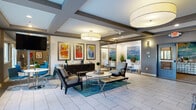This single-floor space serves as a well-appointed leasing office and residential lobby, thoughtfully designed to create a welcoming and professional environment. The main lounge area sets an inviting tone with large-format drum pendant lighting, coffered ceilings with exposed beams, and light-toned large-format tile flooring throughout. A curated seating arrangement — featuring upholstered armchairs, a round white coffee table, and a leather bench — is anchored by a teal area rug, adding a vibrant pop of color. A gallery wall of three bold, colorful framed artworks and a dark console sideboard add sophistication to the lounge. Adjacent to the main seating area, a dedicated business center is enclosed by sleek floor-to-ceiling glass partitions with dark metal framing, housing multiple computer workstations and lush greenery. The entry corridor features textured tile accent walls in warm grey tones, wall-mounted sconces, and locally inspired artwork, including a Tennessee-themed print, lending a regional character to the space. An elevator provides convenient building access. The overall ambiance is modern, polished, and professionally curated, balancing functionality with a warm, residential aesthetic.